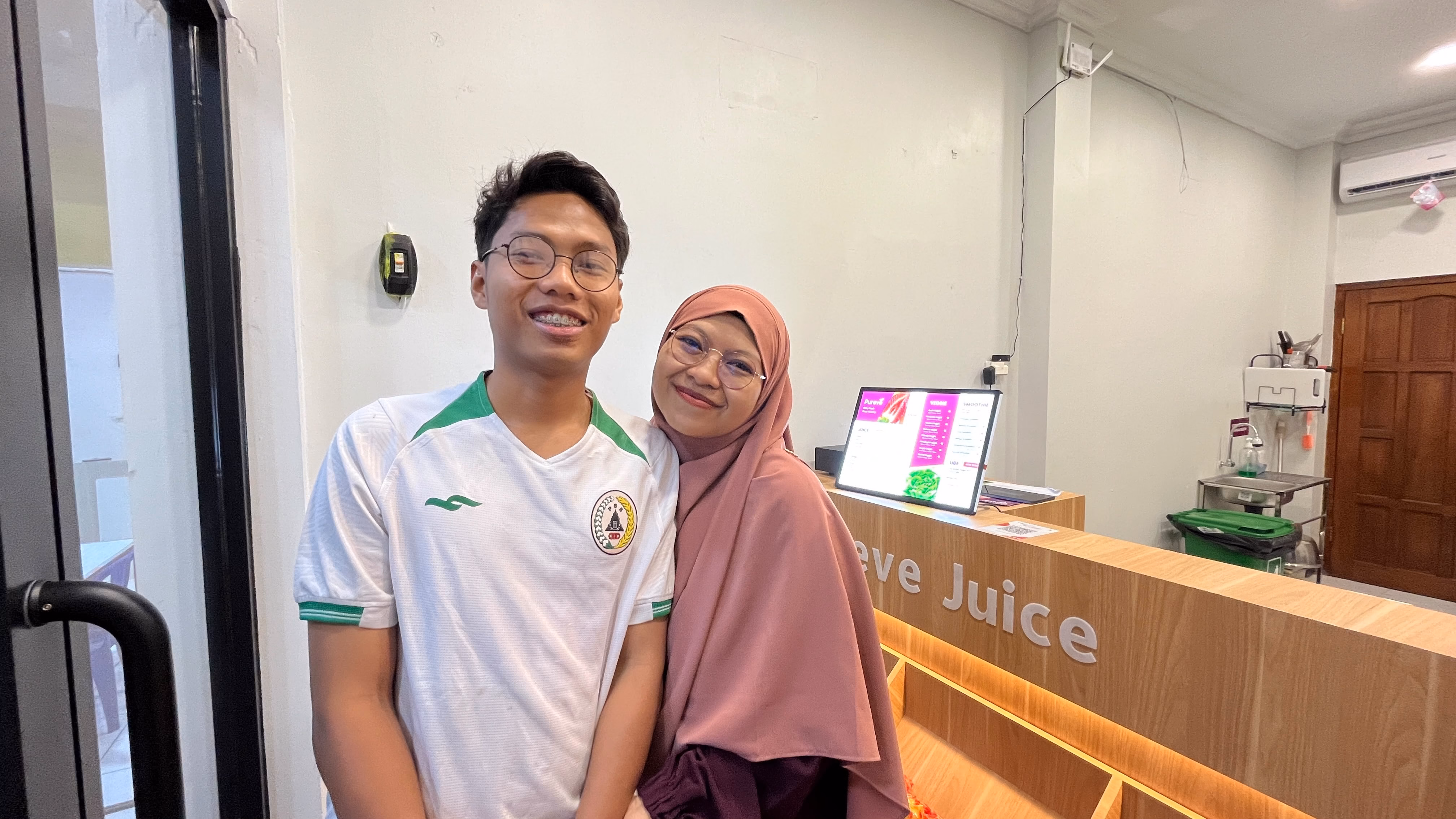 Pria berkacamata mengenakan jersey dan wanita berhijab pink berpose tersenyum di depan meja kasir outlet jus.